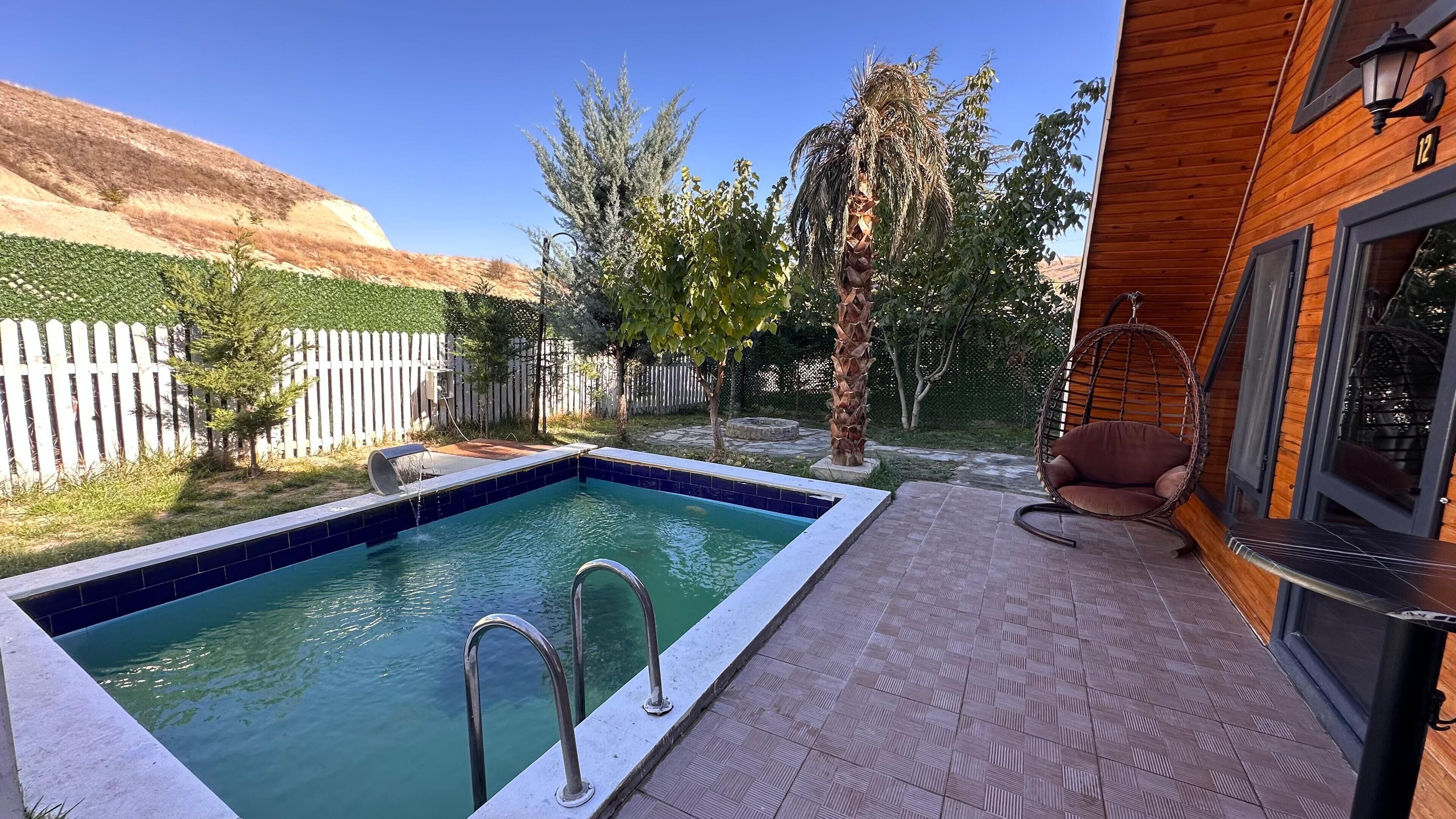Una piscina con cubierta de madera, una silla colgante y un área cercada con árboles.
