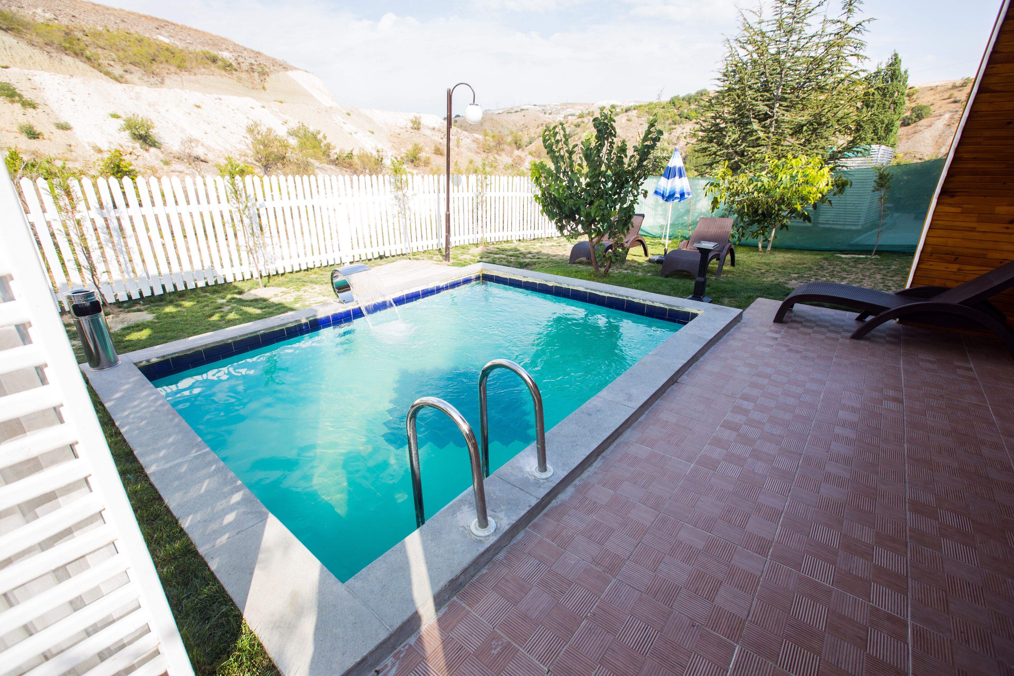 A rectangular swimming pool with metal ladders, surrounded by a white picket fence, a grassy area, and a tiled patio.
