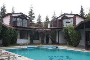 A swimming pool with a geometric pattern on the floor, surrounded by two-story buildings with balconies and a staircase connecting them. - ÇELİK RESORT (Ayas)