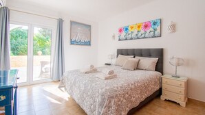 Ein Schlafzimmer mit einem großen Bett, einem Nachttisch, einer Lampe, einem Gemälde und Blick durch eine Glastür.