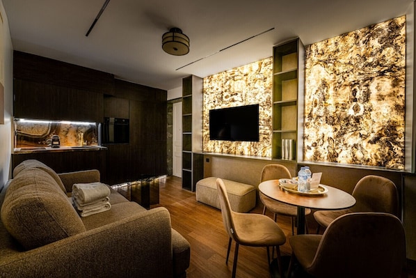 Un salon moderne comprenant un canapé, des fauteuils, une table à manger, un téléviseur et un coin cuisine avec un revêtement mural en marbre.