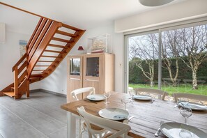 A dining area with a wooden table set for four, a wooden staircase, and a view of the garden through large windows. - Sea view house for 6, garden, beach 400 m away (Sarzeau)
