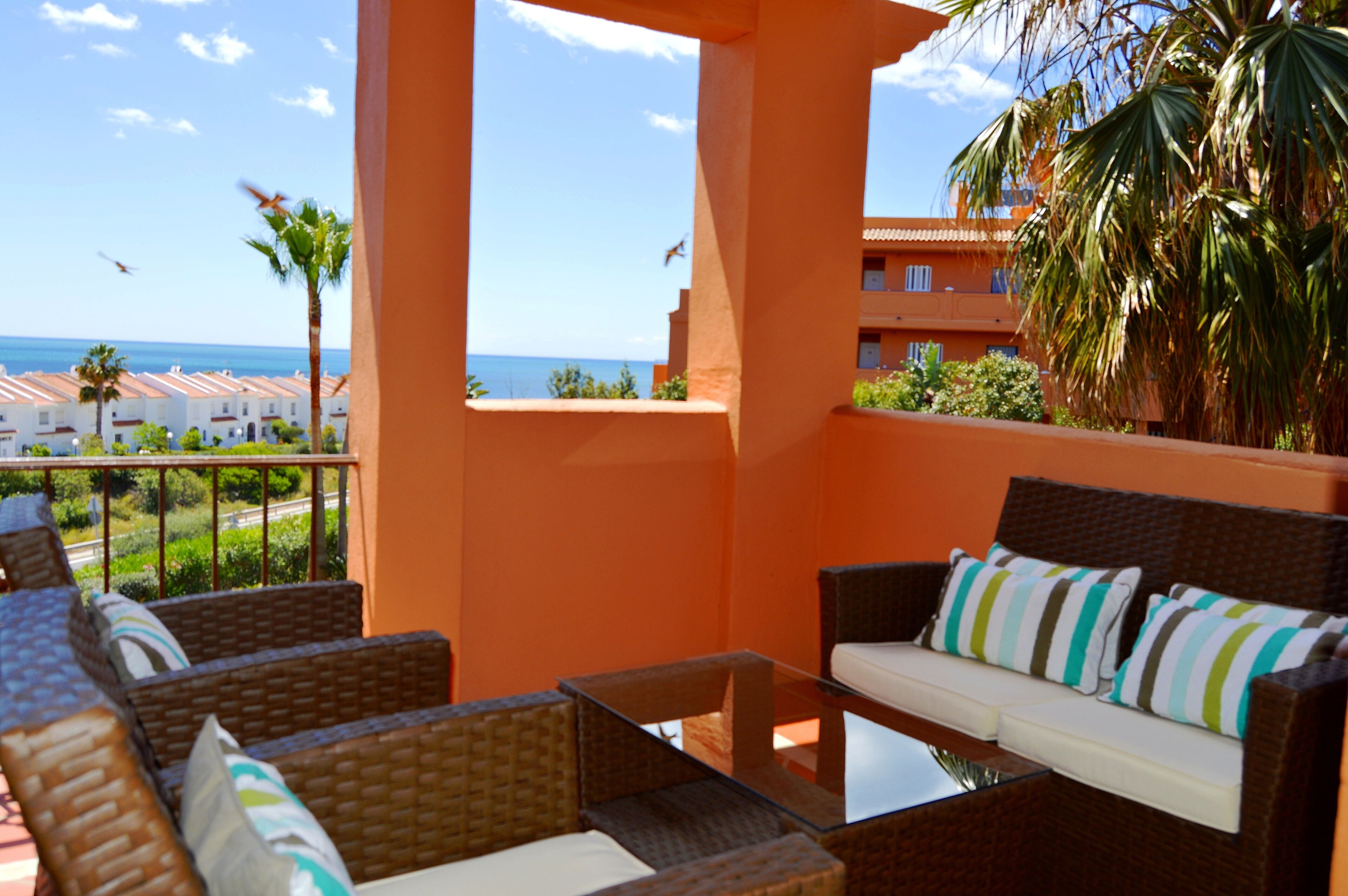 A balcony with wicker furniture, a glass table, and a view of the ocean and buildings.