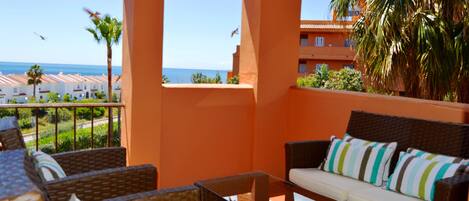A balcony with wicker furniture, a glass table, and a view of the ocean and buildings.