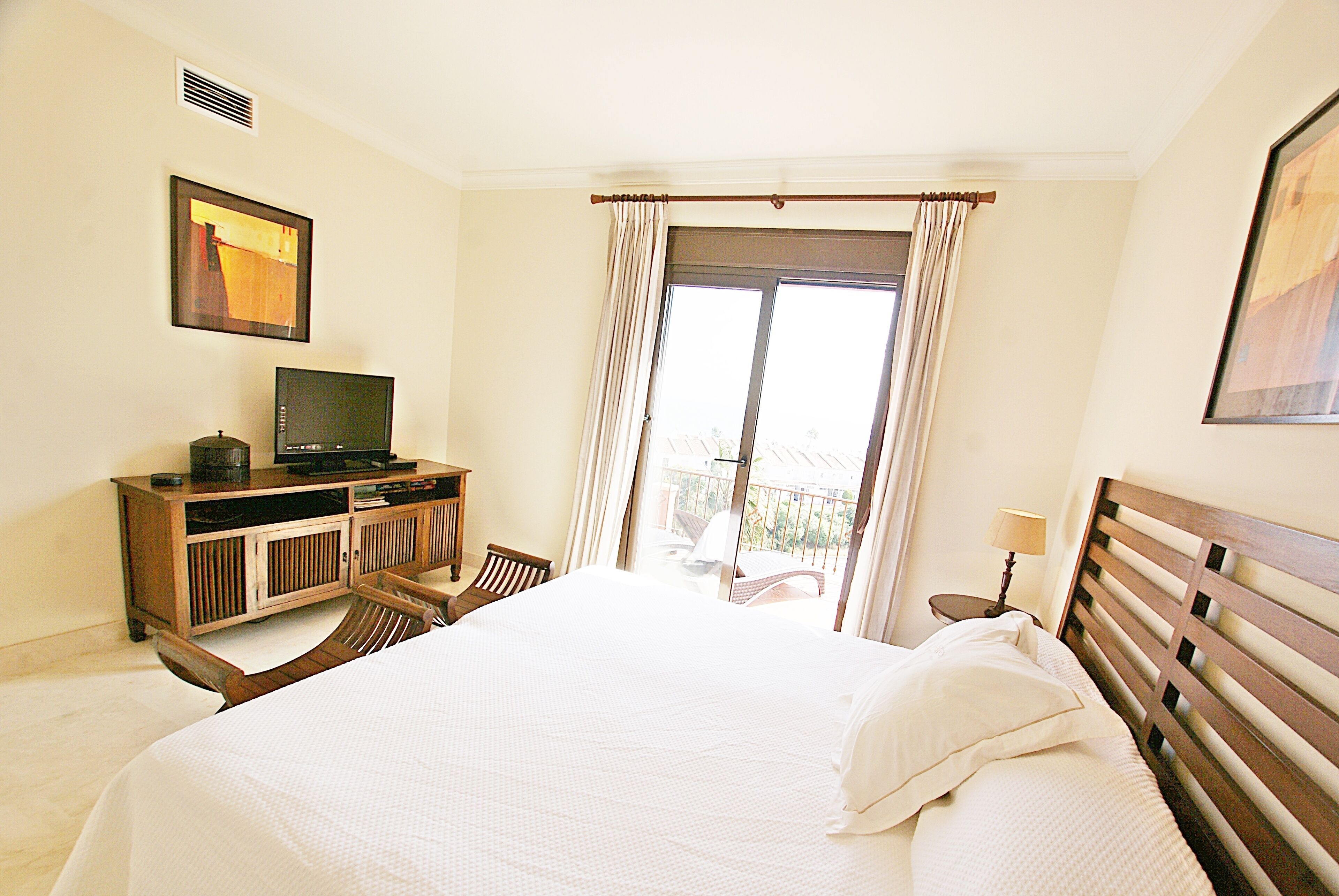 A hotel room with a bed, a TV on a wooden cabinet, a chair, a balcony view, and a painting on the wall.