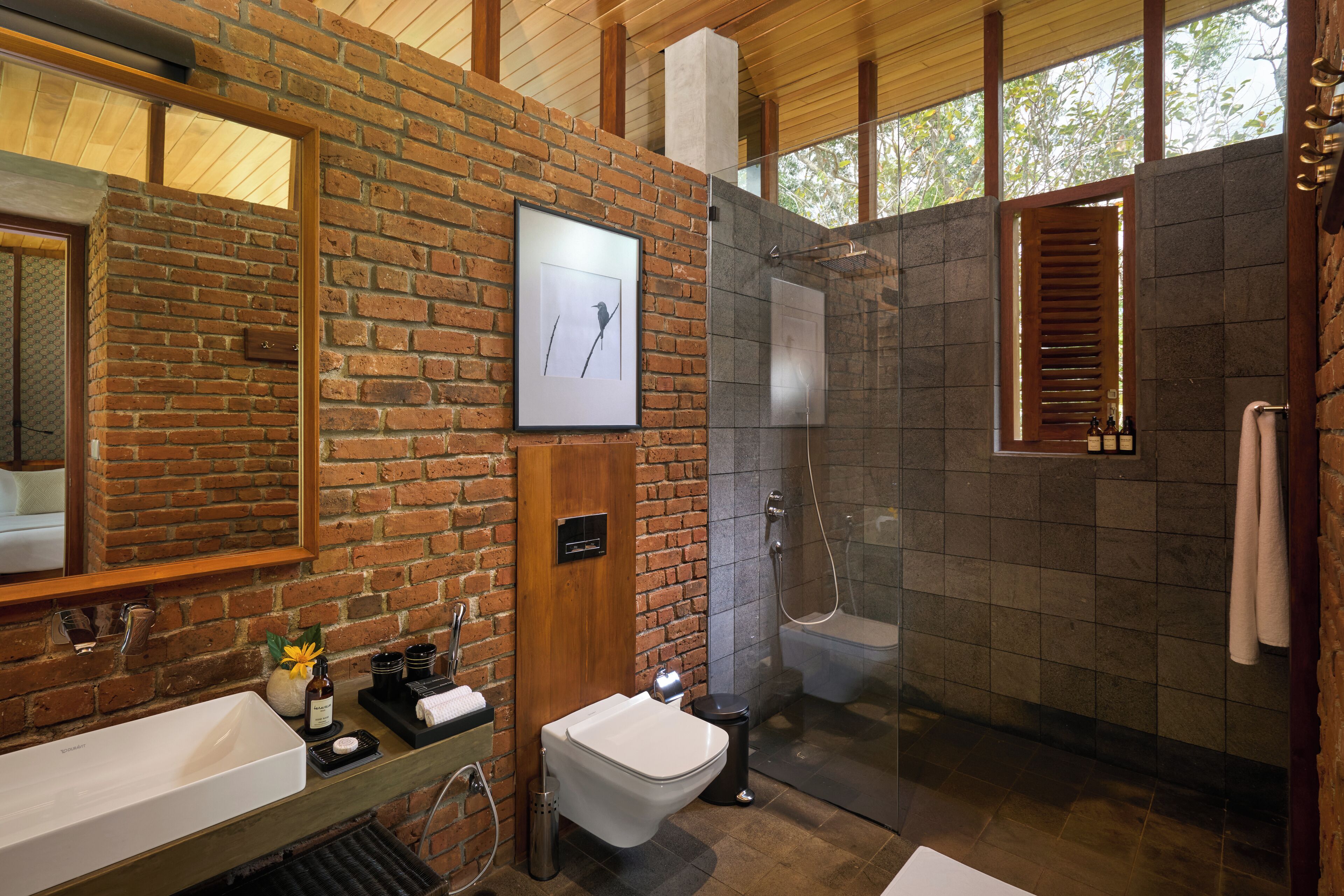 Ein modernes Badezimmer mit Backsteinwand, großem Spiegel, weißem Waschbecken, Toilette und einer Duschkabine.