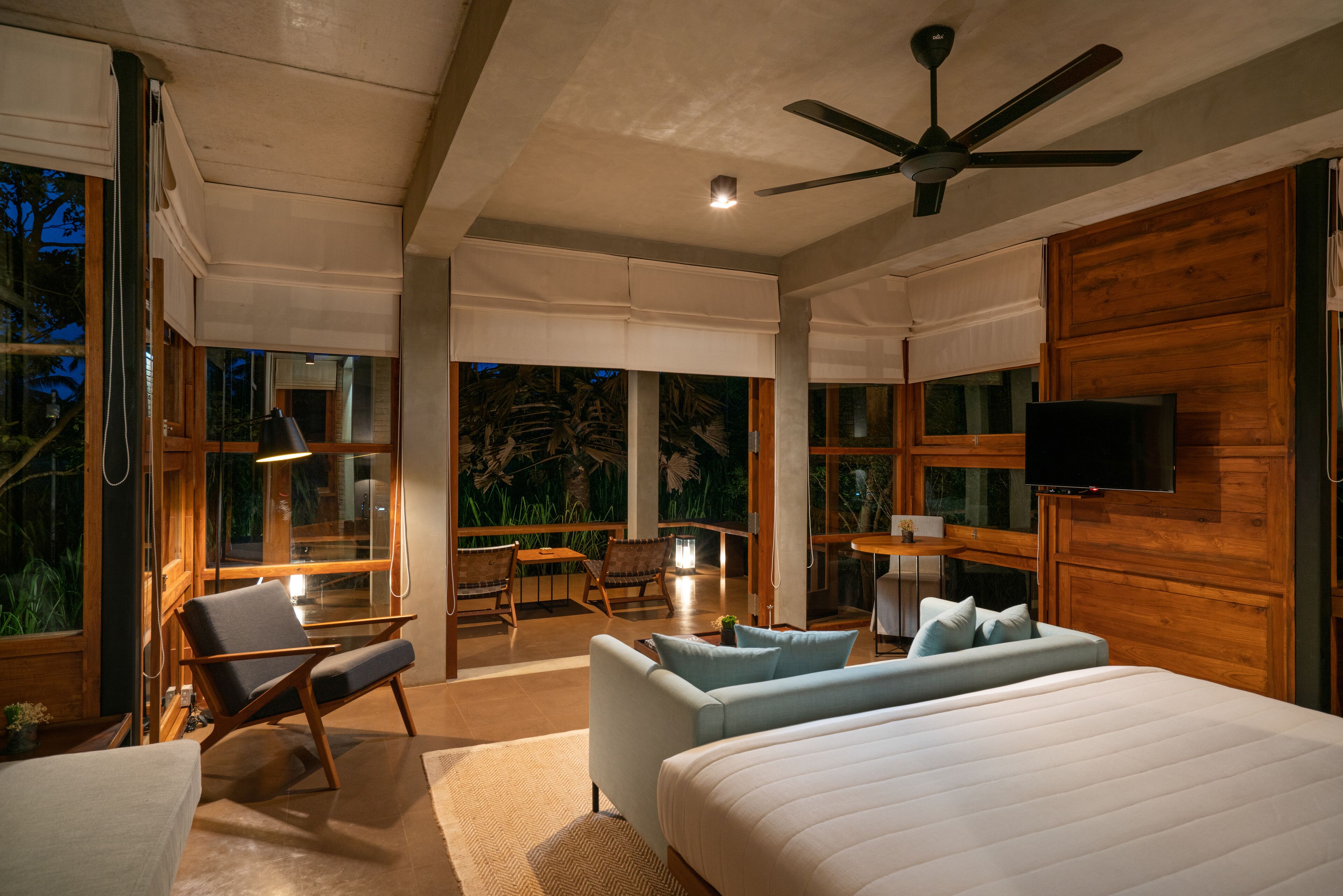 Une chambre moderne avec un grand lit, un canapé et un fauteuil. On y trouve un ventilateur de plafond, une télévision et de grandes fenêtres donnant sur l’extérieur.