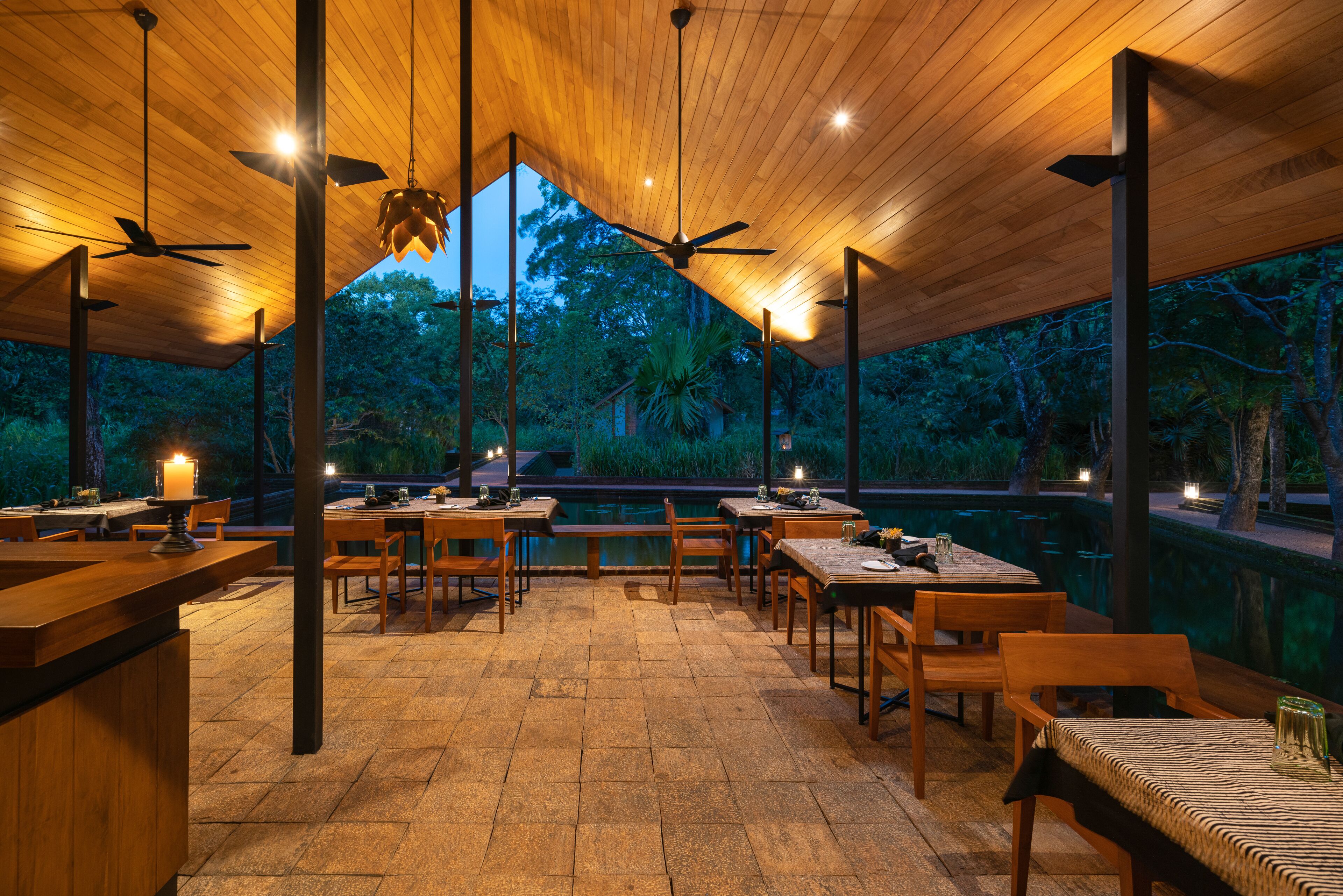 Un espacio al aire libre para comer con mesas y sillas de madera, ventiladores de techo y vistas a árboles.