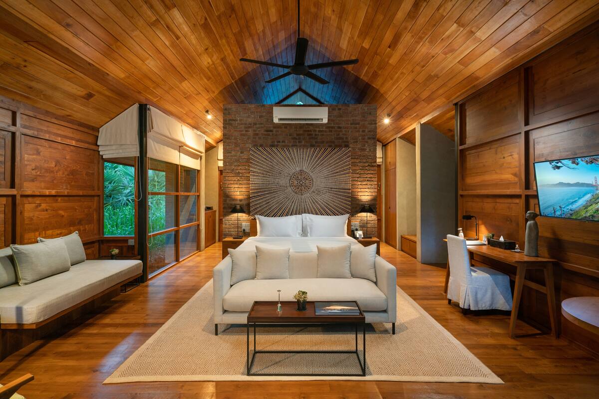 Une chambre moderne avec un grand lit, un canapé blanc, un ventilateur de plafond en bois et une grande fenêtre donnant sur un paysage verdoyant.