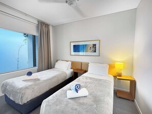A hotel room with two beds, a window with curtains, a framed artwork, a bedside lamp, and a small table.