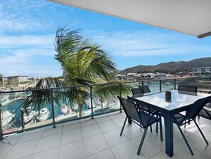 A balcony with a glass railing overlooking a marina with boats, a palm tree, and a view of buildings across the water. - Blue on Blue Apartment 932 (Nelly Bay)