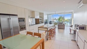 A modern kitchen with a glass dining table, stainless steel appliances, and a view of palm trees through large windows. - Blue on Blue Apartment 932 (Nelly Bay)
