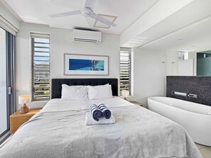Une chambre d’hôtel moderne avec un grand lit, un ventilateur de plafond, un climatiseur fixé au mur, un tableau encadré accroché au mur et une vue sur l’extérieur par la fenêtre.