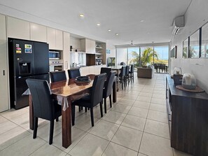 A modern kitchen with a dining area, featuring a large island, dark chairs, and a view of the outdoors. - Blue on Blue Apartment 932 (Nelly Bay)
