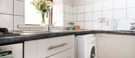 A kitchen with white tiles, a washing machine, a digital clock, and a window with a plant.