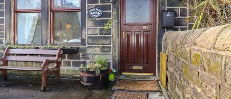 A stone cottage with a wooden door marked "183", a bench, and potted plants.