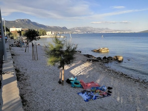 A pebble beach with a small tree, a blue and red cloth, and a rocky outcrop in the water. - Apartments Vujina (Kastela)