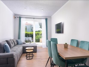 A modern living room with a grey sofa, teal chairs, a wooden dining table, and a TV on the wall. - Apartments Vujina (Kastela)