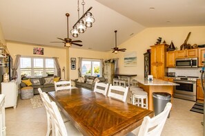 A spacious living area with a wooden dining table, white chairs, a sofa, and a kitchen area with wooden cabinets and appliances.