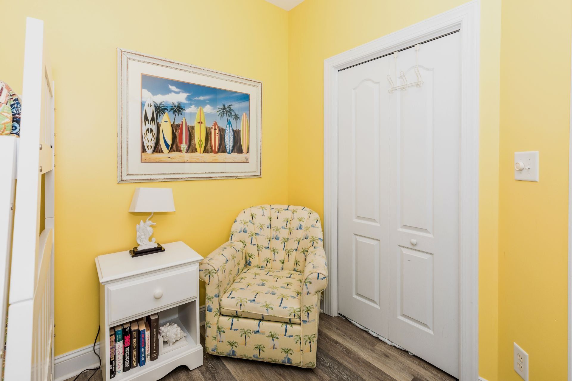 Une pièce avec un mur jaune, un tableau encadré représentant des planches de surf, une porte blanche, une petite table de chevet blanche avec une lampe et un fauteuil à motifs.