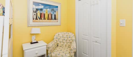 Ein Zimmer mit gelber Wand, einem gerahmten Bild von Surfbrettern, einer weißen Tür, einem kleinen weißen Nachttisch mit Lampe und einem gemusterten Sessel.
