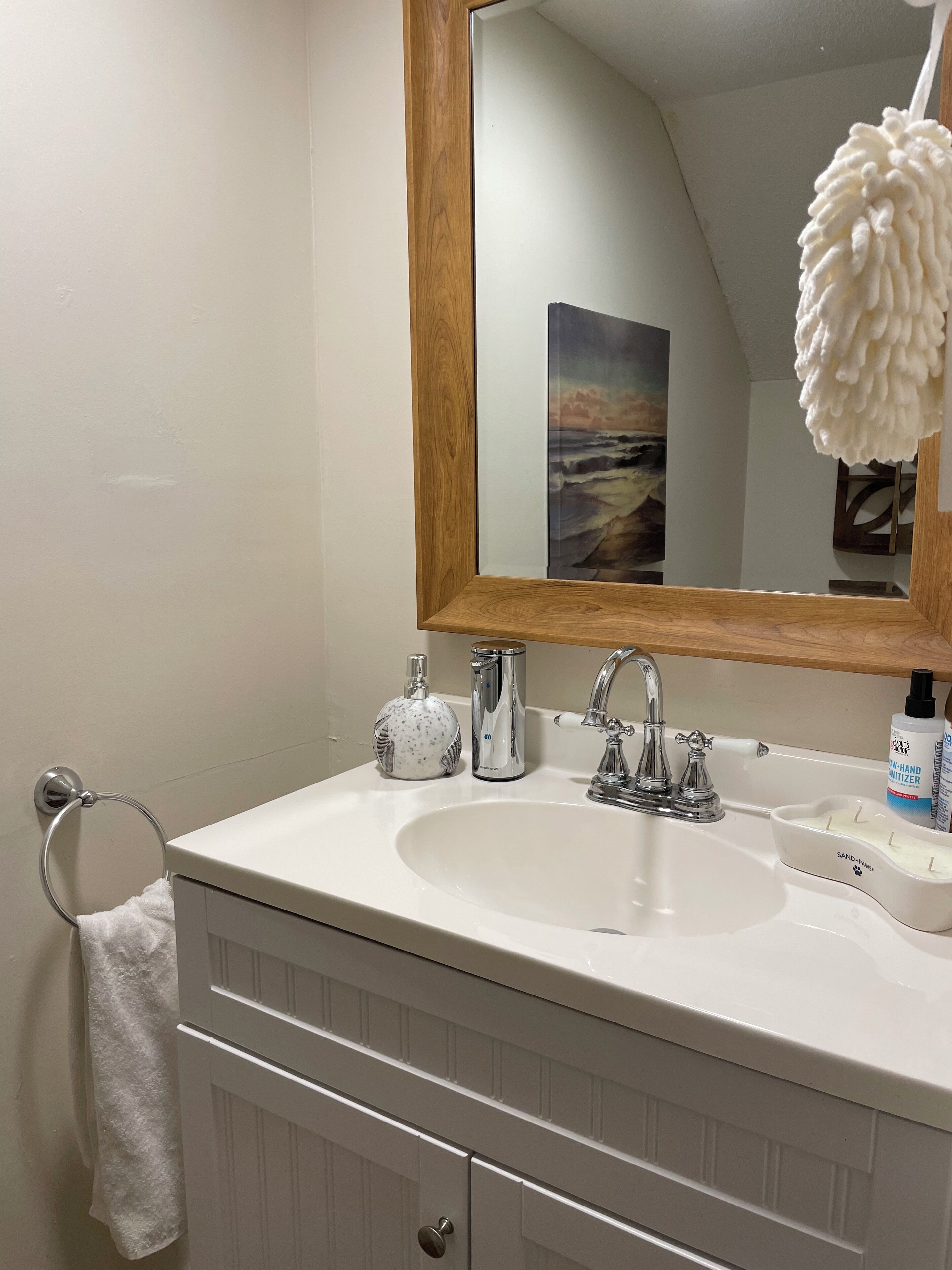 Une salle de bain avec un lavabo blanc, un miroir encadré en bois, un porte-serviettes, un distributeur de savon, un flacon de gel hydroalcoolique et un porte-bougie.