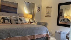 A bedroom with a large bed, a bedside table, a chair, a small table, a mirror, and a wall with a picture and a sign.