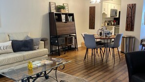 A living room with a sofa, coffee table, and a dining area with chairs and a table. There is a ceiling fan and a kitchen in the background.