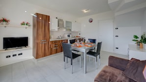 A modern kitchen with white cabinets, a dining table with chairs, a TV mounted on the wall, and a brown sofa in the living area.