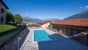 A rectangular swimming pool with clear water, surrounded by stone walls and a stone floor, set against a backdrop of mountains and traditional buildings with red-tiled roofs.