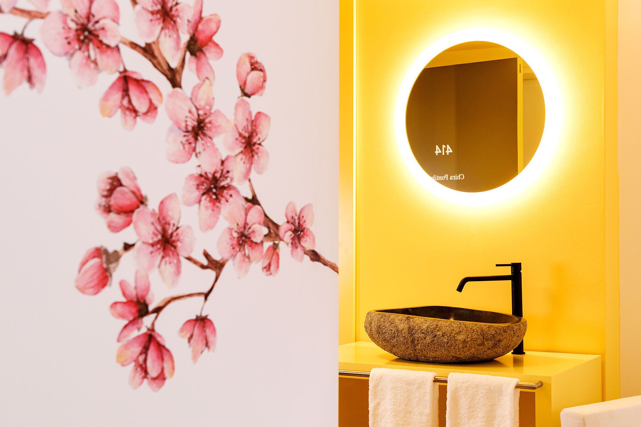 A bathroom with a stone sink, a round mirror, and a wall with cherry blossom artwork.