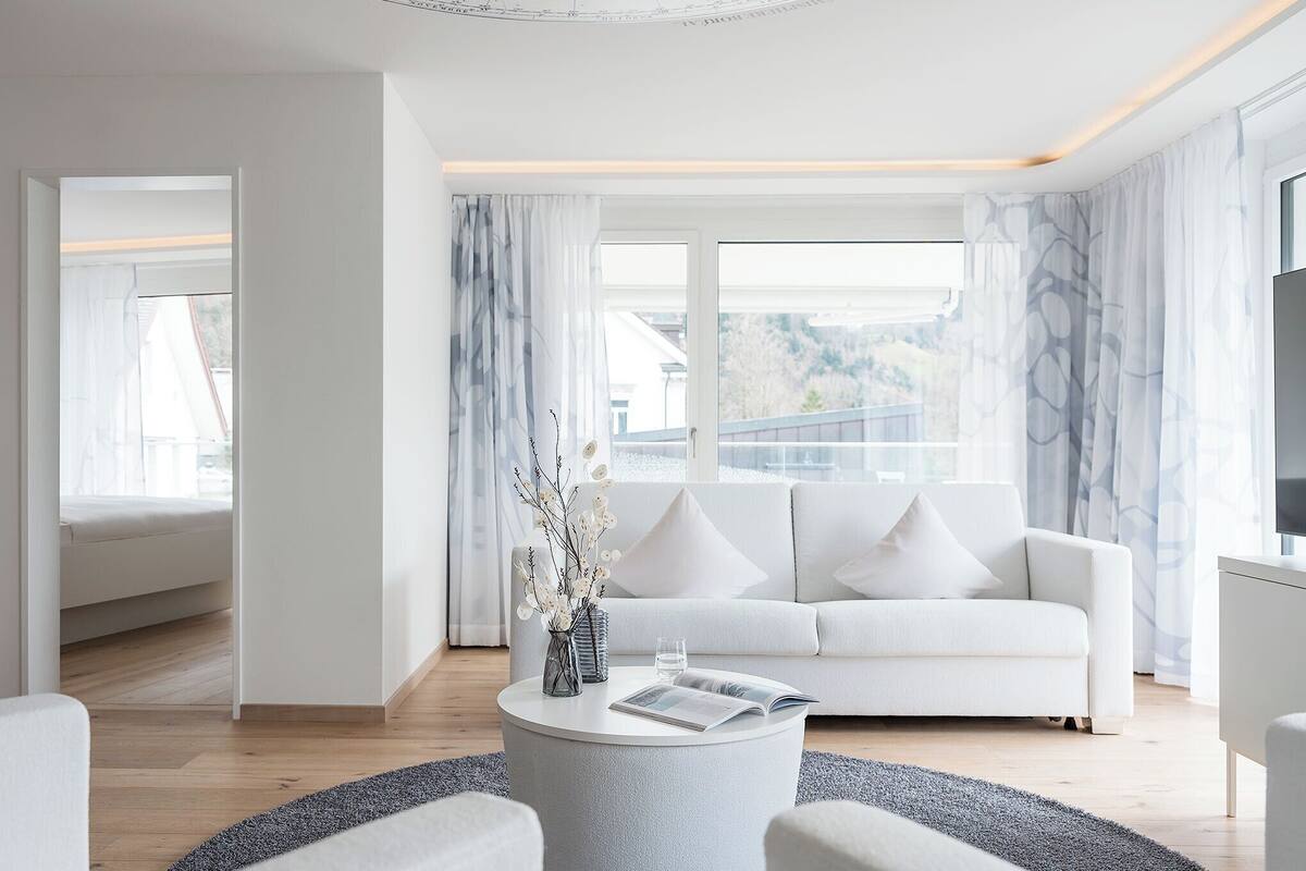 A modern living room with a white sofa, a round coffee table with a vase and book, and large windows with curtains.