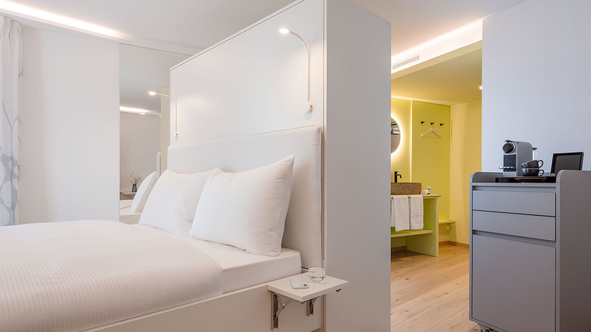Een moderne slaapkamer met een groot bed, nachtkastje en uitzicht op een badkamer met wastafel en spiegel.