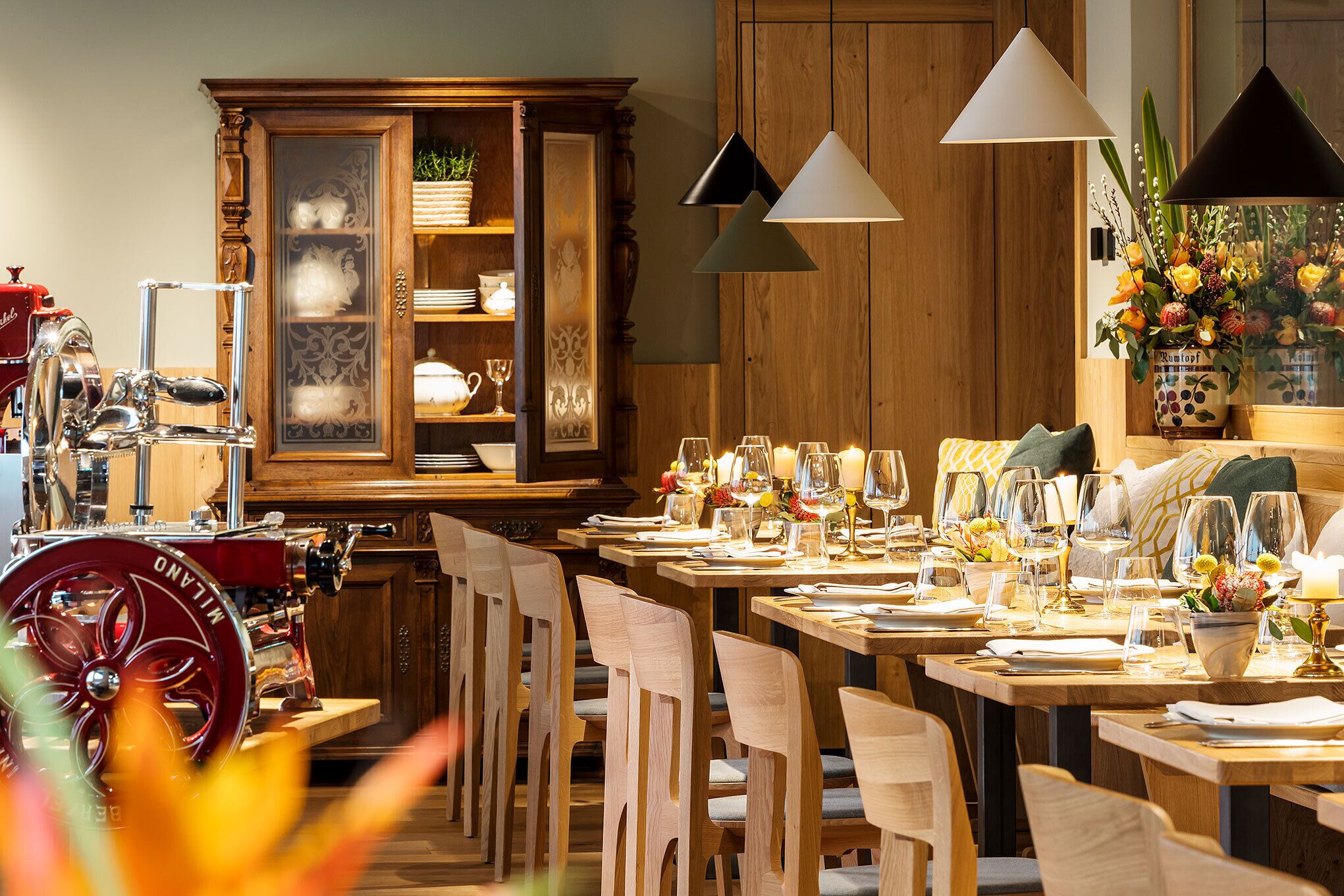 Un restaurante con mesas y sillas de madera, un aparador grande con puertas de cristal y lámparas colgantes.