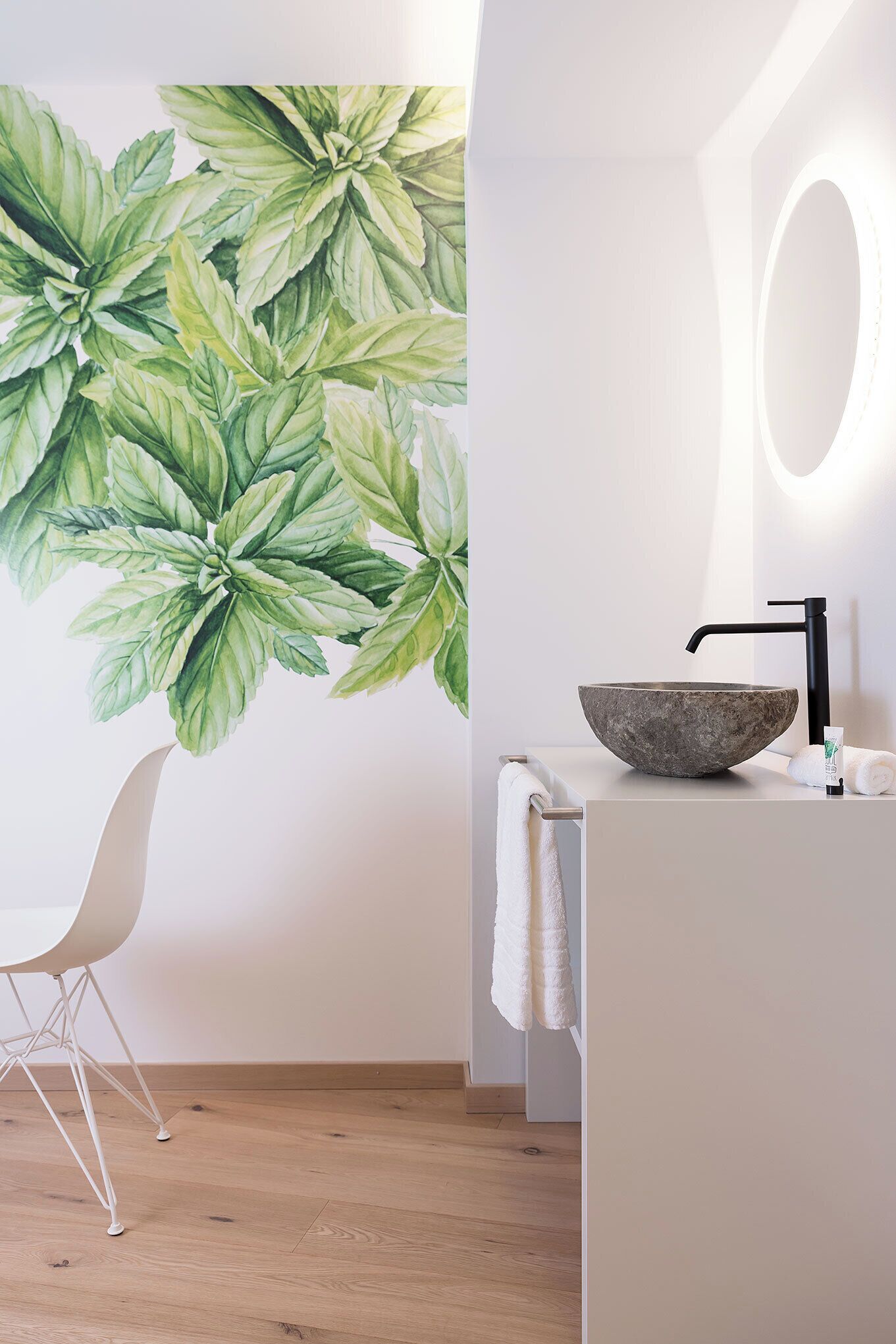 A bathroom with a large wall mural of green leaves, a white sink with a stone basin, a black faucet, and a white chair.