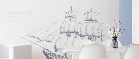 Una sala de estar moderna con un gran mural de un barco velero, muebles blancos y un jarrón con ramas.