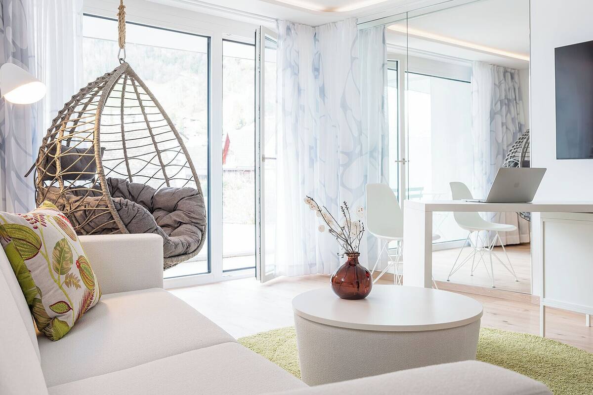 A modern living room with a hanging chair, a sofa with decorative pillows, a round coffee table, and a laptop on a desk.
