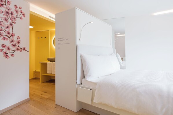 Un dormitorio moderno con una cama blanca, una mesita de noche empotrada y un espejo montado en la pared.