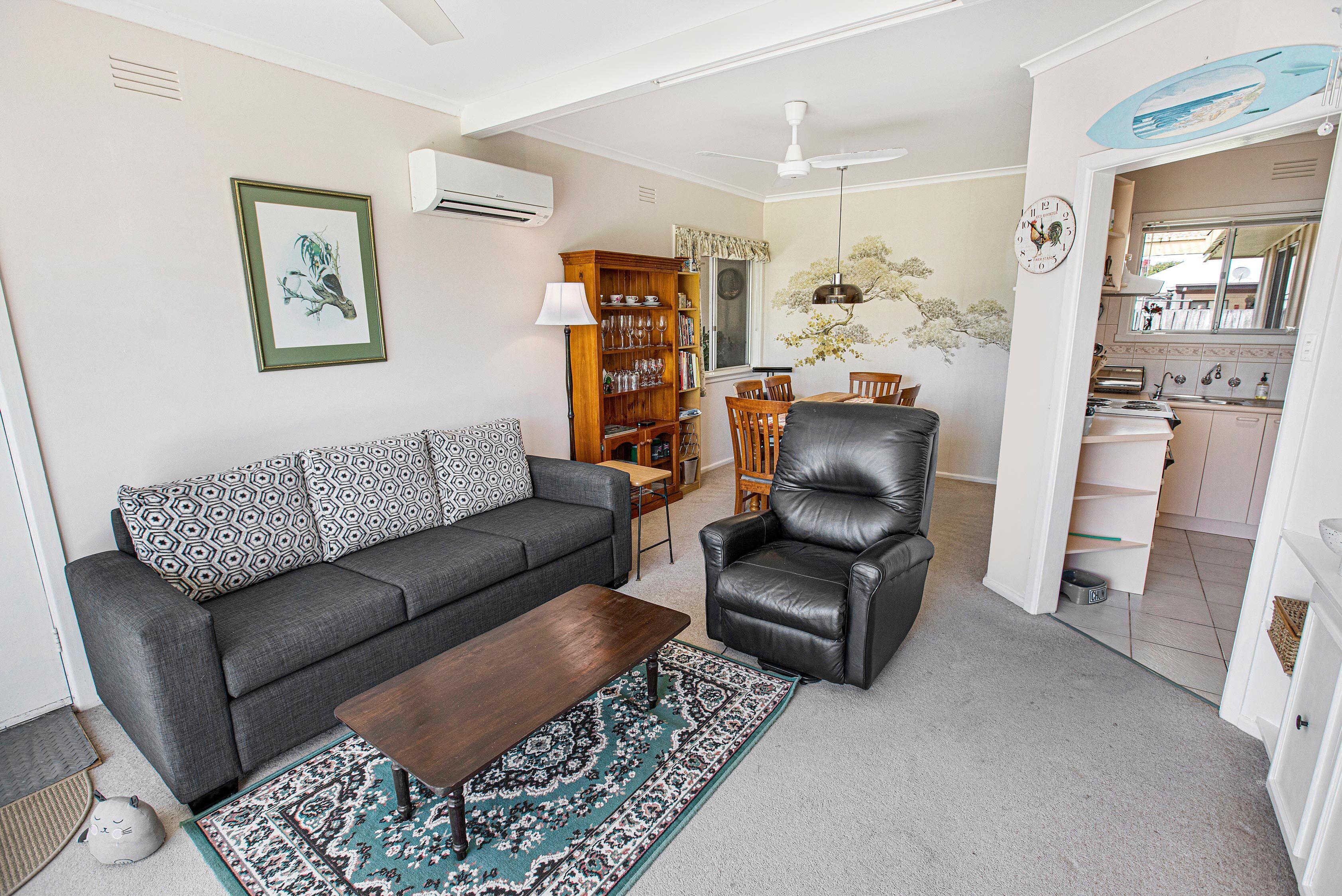 A living room with a sofa, armchair, coffee table, and a rug. There is a bookshelf, a dining table with chairs, and a kitchen area visible through an open door.