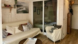 Ein gemütliches Wohnzimmer mit einer Couch, einem Sessel und einem kleinen Tisch. Eine Glastür verschiebt sich zu einem Schlafzimmer.