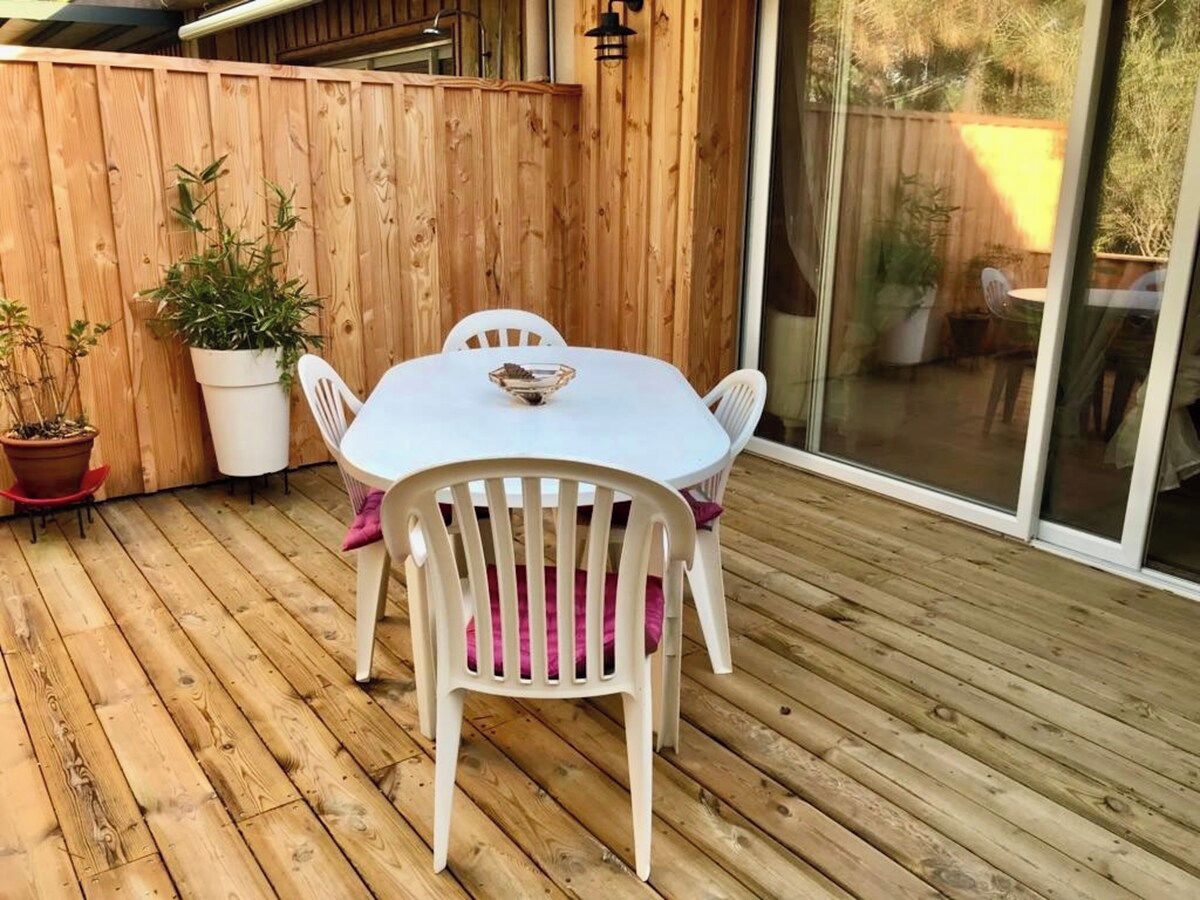 Une terrasse en bois avec une table et des chaises blanches, des plantes en pot et une porte-fenêtre coulissante.
