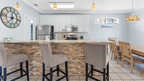 A modern kitchen with white cabinets, a central island with bar stools, a dining area with a wooden table and chairs, and pendant lighting. - Pet Friendly ~ Bunk Room ~ Community Pool (Miramar Beach)