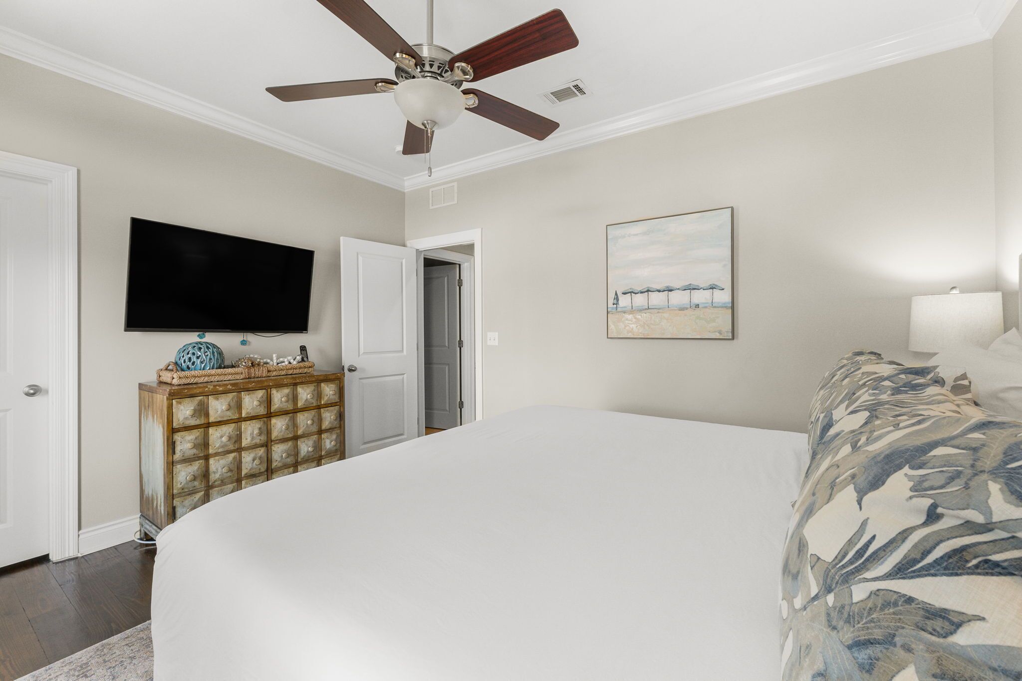 A bedroom with a bed, a ceiling fan, a television, a dresser, and a painting on the wall.