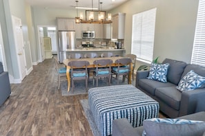 A modern kitchen with stainless steel appliances, a dining area with wooden chairs and a table, and a grey sofa with patterned cushions.