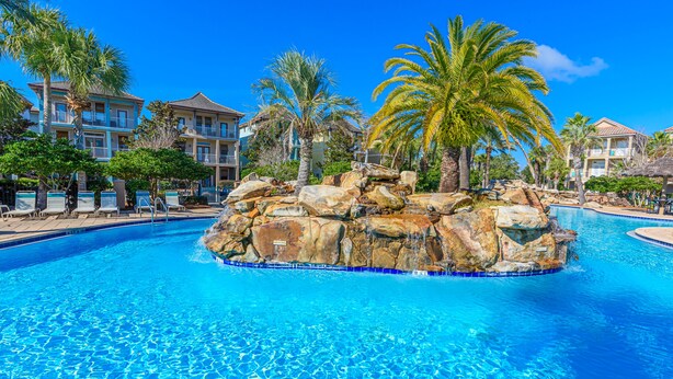 Une piscine à l’eau d’un bleu cristallin, entourée de rochers et de palmiers, avec un bâtiment en arrière-plan.
