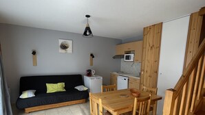 Een knus woongedeelte met een houten eettafel, een zwarte bank, een kitchenette en een trap.