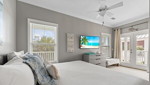 Une chambre avec un grand lit, un ventilateur de plafond, une télévision et une vue sur la plage par la fenêtre.