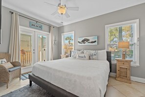 Een slaapkamer met een groot bed, een plafondventilator, een raam met luiken en een deur met een bordje 'BEACH'.