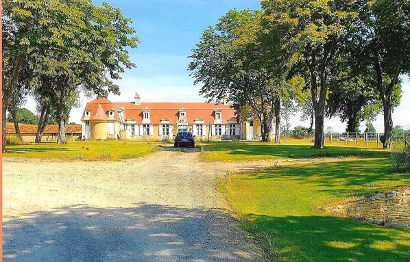 A large building with a red roof and multiple windows, surrounded by trees and a gravel path.