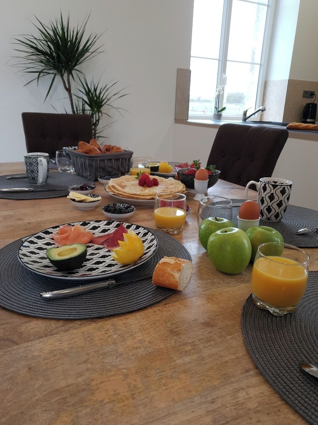Una mesa de desayuno con platos de comida, vasos de zumo de naranja y una planta al fondo.